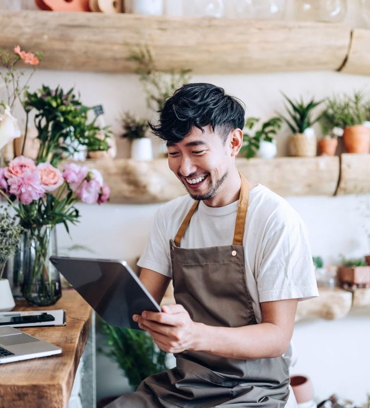 Happy business owner reviewing big savings and lower processing fees on a tablet in a flower shop, excited about other modern solutions from Virtue Payments who provide amazing support services