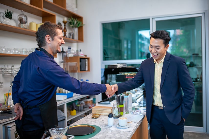 Business advisor meeting with café owner, shaking hands over counter to celebrate partnership and personalized support from Virtue Payments
